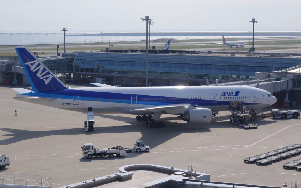 On the ramp at Tokyo-Haneda, an ANA Boeing 777-200 with a pod-loader, belt loader, & towbar tractor by the plane, and a lavatory service truck going by.